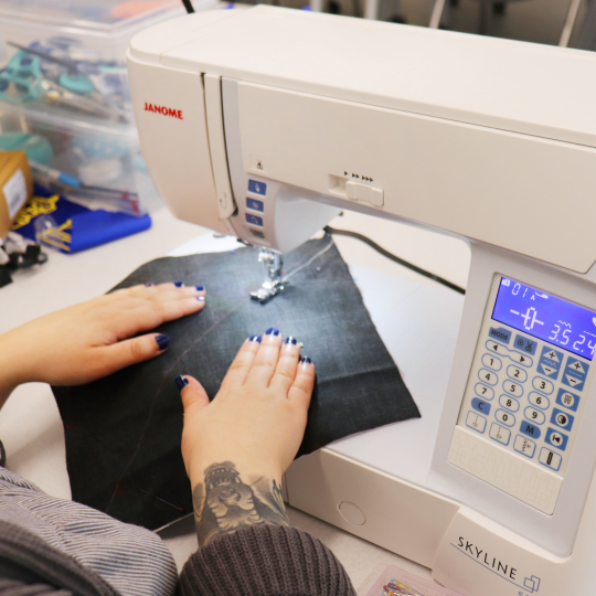 A person sews a black square of fabric on a white sewing machine at a table in the Makerspace at McLean Branch.