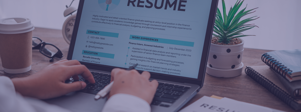 A person types a resume on a laptop at a table. There is a plant beside the laptop.