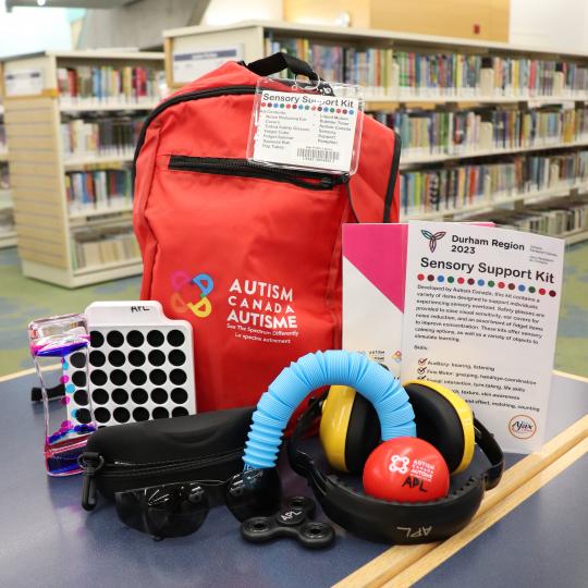 A red backpack on a table at Main Branch with a variety of items such as a fidget spinner, sunglasses and noise-cancelling headphones.